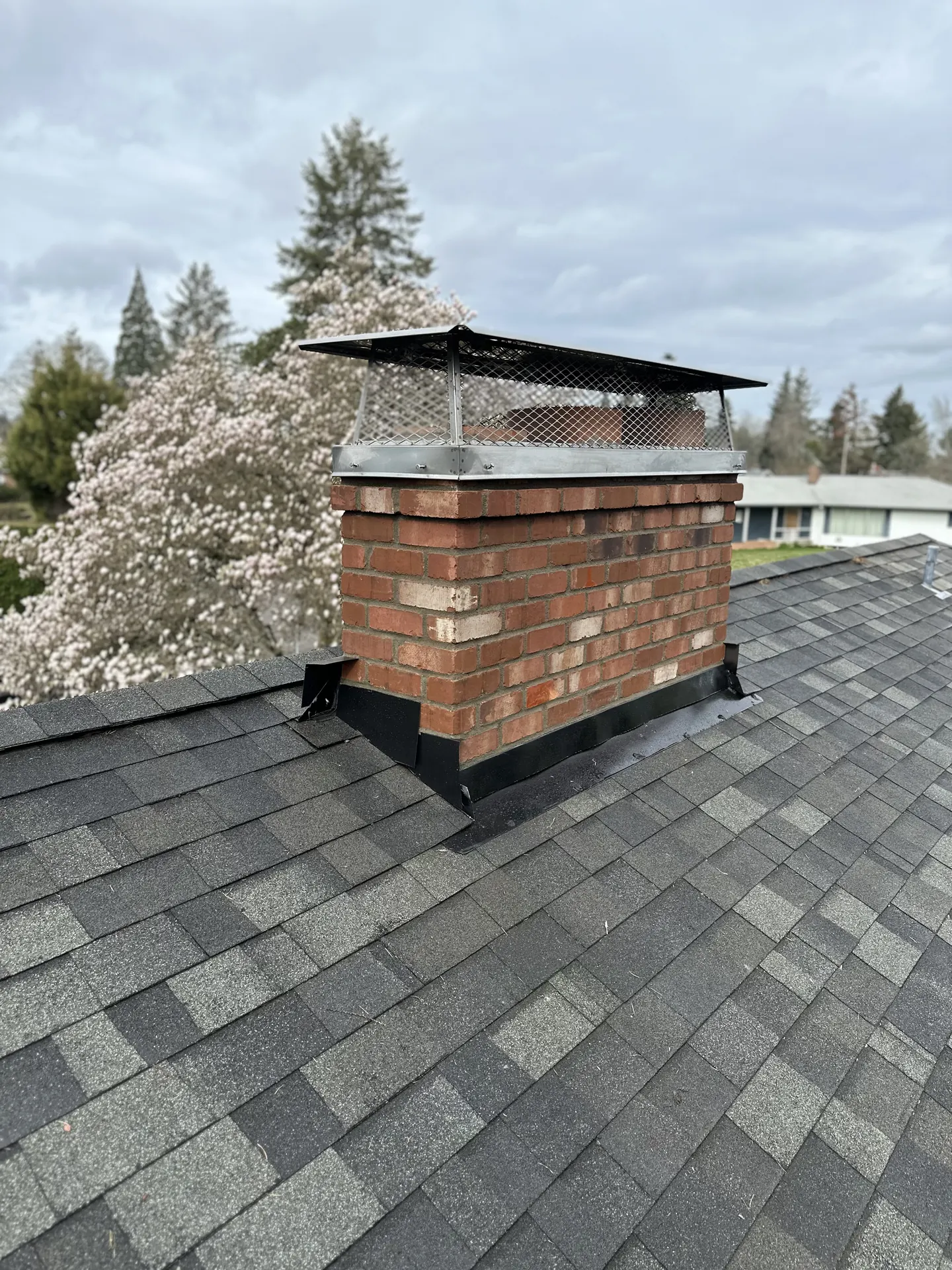A wide custom black chimney cap on a rebuilt brick chimney against spring blossoms